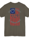 rear view of a army green colored fabric shirt with americantees.us logo on front and betsy ross flag with the phrase give me liberty or give me death and 1776 on the back