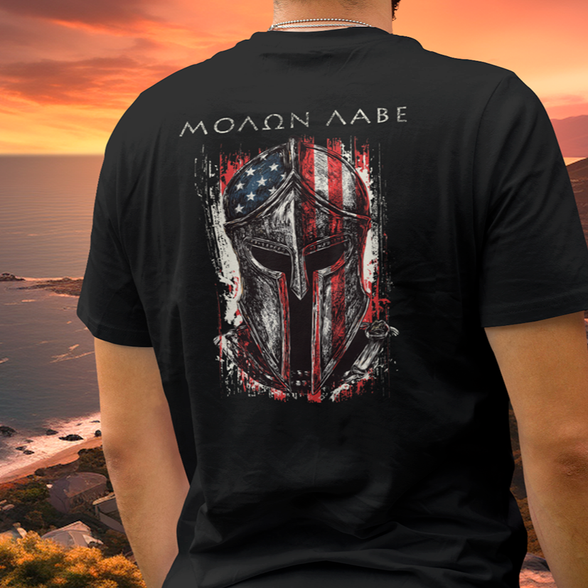 man overlooking the beach at sunset wearing a black color fabric tee with americantees.us logo on front with spartan helmet covered with american flag and motto molon labe patriotic shirt