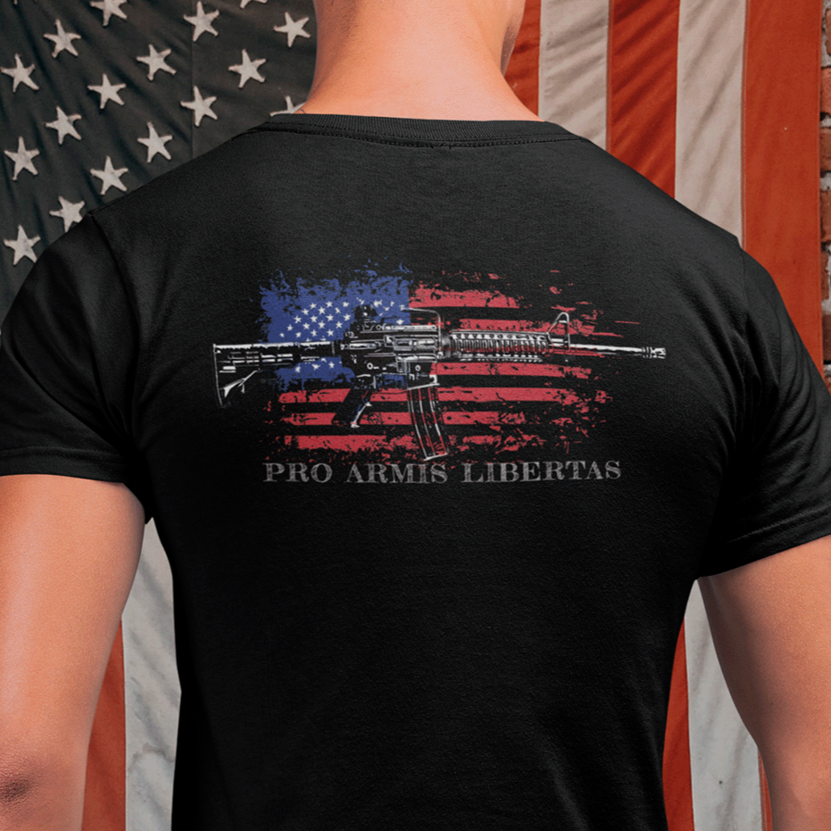 Man standing in front of an American flag wearing a Black color fabric patriotic shirt rear view has an americantees.us logo with eagle head on front , rear view has distressed American flag with modern sporting rifle with the log Pro Armis Libertas meaning For Arms, Freedom