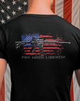 Man standing in front of an American flag wearing a Black color fabric patriotic shirt rear view has an americantees.us logo with eagle head on front , rear view has distressed American flag with modern sporting rifle with the log Pro Armis Libertas meaning For Arms, Freedom