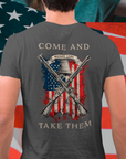 rear view of a mean standing in front of a USA flag wearing a patriotic shirt white color fabric tee with logo on front and american flag with liberty bell and crossed rifles on back, motto that states Come and Take Them and molon labe on scroll