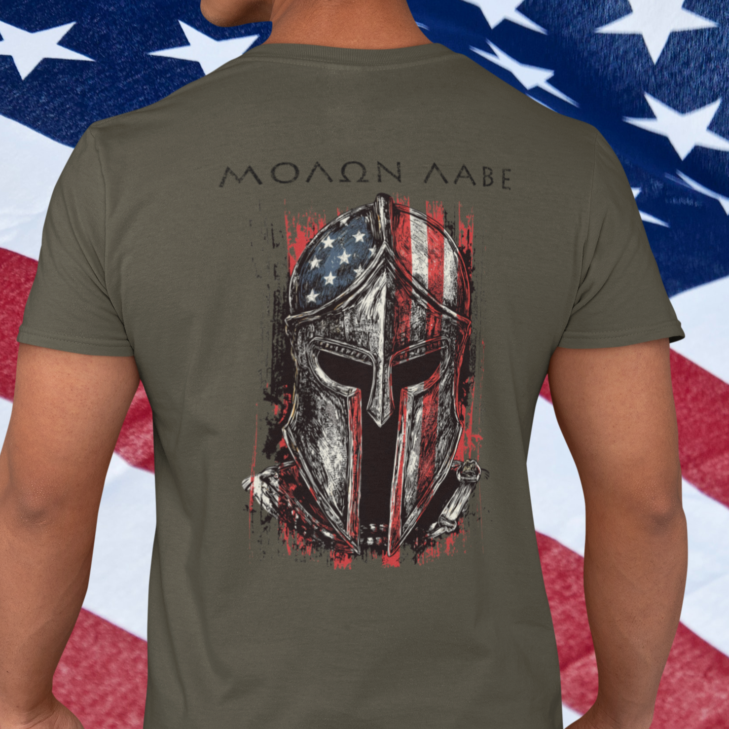 man standing in front of a USA flag rear view army green color fabric tee with americantees.us logo on front with spartan helmet covered with american flag and motto molon labe patriotic shirt