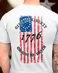 rear view of a a man wearing a white colored fabric shirt with americantees.us logo on front and betsy ross flag with the phrase give me liberty or give me death and 1776 on the back