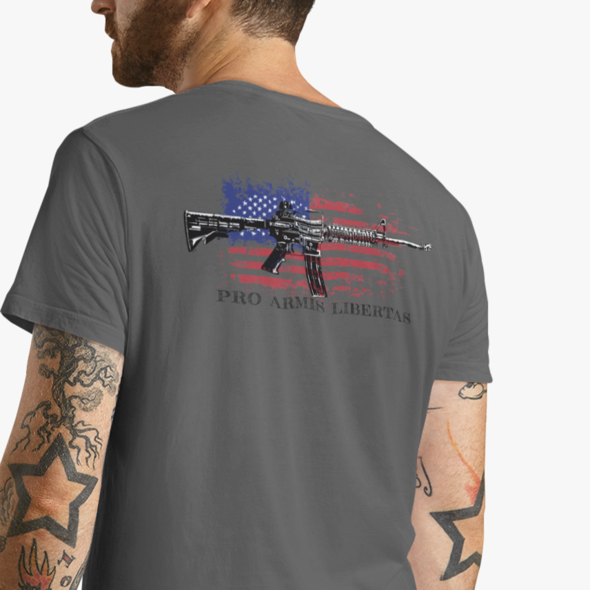 Man wearing an Asphalt color fabric patriotic shirt rear view has an americantees.us logo with eagle head on front , rear view has distressed American flag with modern sporting rifle with the log Pro Armis Libertas meaning For Arms, Freedom