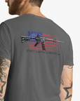 Man wearing an Asphalt color fabric patriotic shirt rear view has an americantees.us logo with eagle head on front , rear view has distressed American flag with modern sporting rifle with the log Pro Armis Libertas meaning For Arms, Freedom