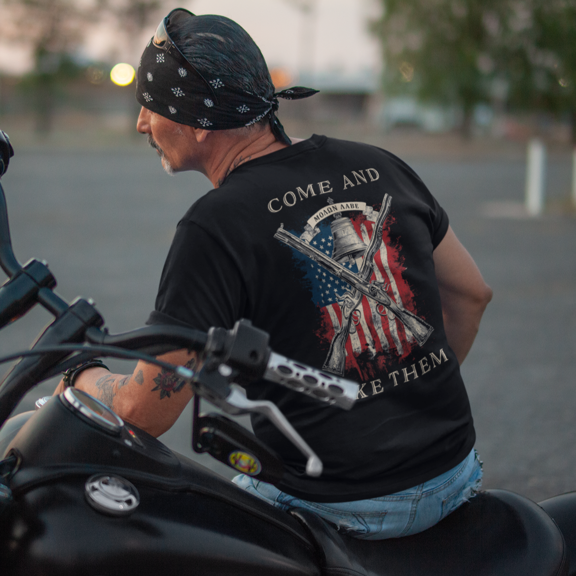 rear view of a man on a motorcycle wearing a patriotic shirt white color fabric tee with logo on front and american flag with liberty  bell and crossed rifles on back, motto that states Come and Take Them  and molon labe on scroll
