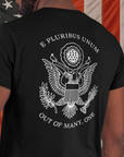 rear view of a man standing in front of an American flag wearing a black colored fabric patriotic shirt with an americantees.us logo on front and the great seal of the united states on the back
