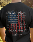 rear view of a man standing in a field wearing black fabric colored patriotic shirt with American flag background and the preamble of the united states constitution, fronto fo shirt has americantees.us logo