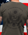 rear view of a man standing in front of an American flag wearing a army green colored fabric patriotic shirt with an americantees.us logo on front and the great seal of the united states on the back