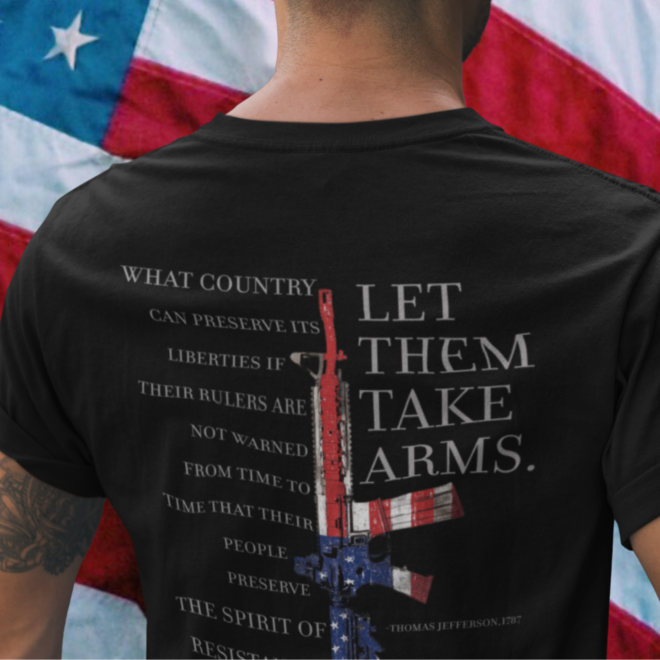 rear view of a man standing in front of an American flag wearing a black color fabric patriotic shirt with americantees.us logo on front and Thomas Jefferson quote on back.