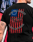 rear view of a man with tattoos and wearing a black colored fabric shirt with americantees.us logo on front and betsy ross flag with the phrase give me liberty or give me death and 1776 on the back