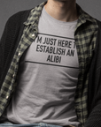 im-just-here-to-establish-an-alibi-athletic-heather-t-shirt-bella-canvas-t-shirt-mockup-featuring-a-serious-man-sitting-on-a-stool