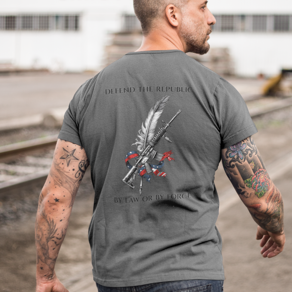Patriotic man wearing an asphalt colored t-shirt with a graphic design of a rifle  crossed with a quill fountain pen and an American flag ribbon, with the quote Defend the Republic by law or by force. 