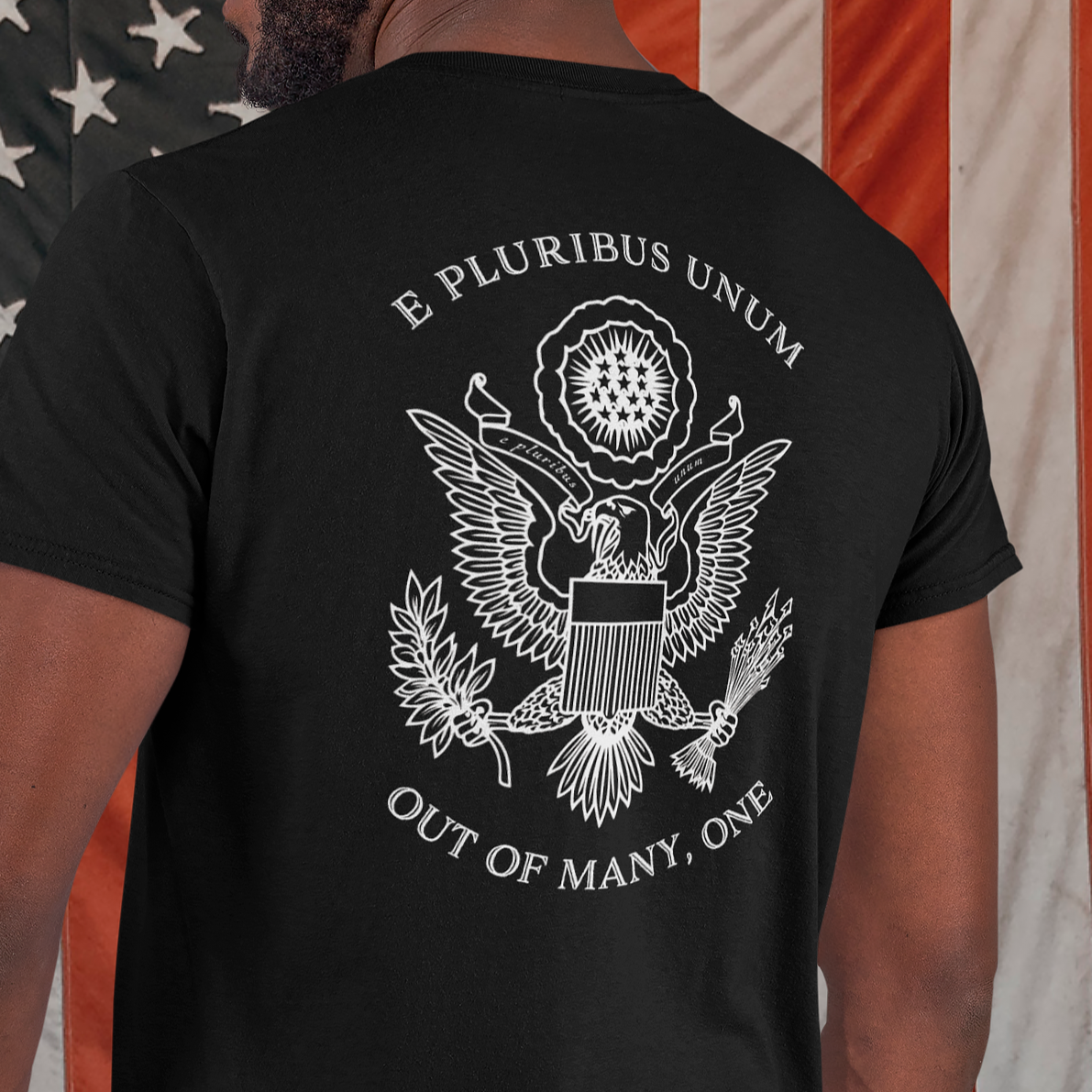 rear view of a man standing in front of an American flag wearing a black colored fabric patriotic shirt with an americantees.us logo on front and the great seal of the united states on the back 