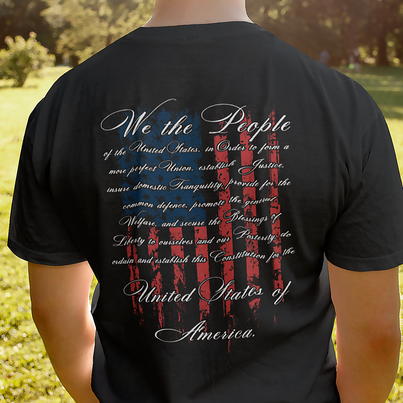 rear view of a man standing in a field wearing black fabric colored patriotic shirt with American flag background and the preamble of the united states constitution, fronto fo shirt has americantees.us logo