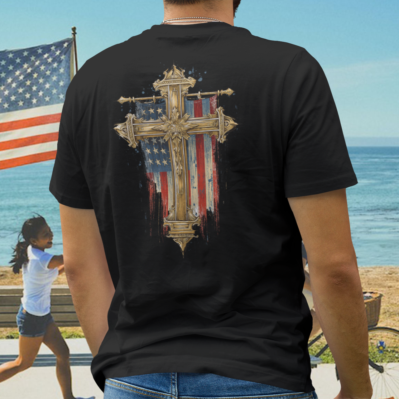 Person wearing a black t-shirt with a cross and American flag design, standing on a beach.
