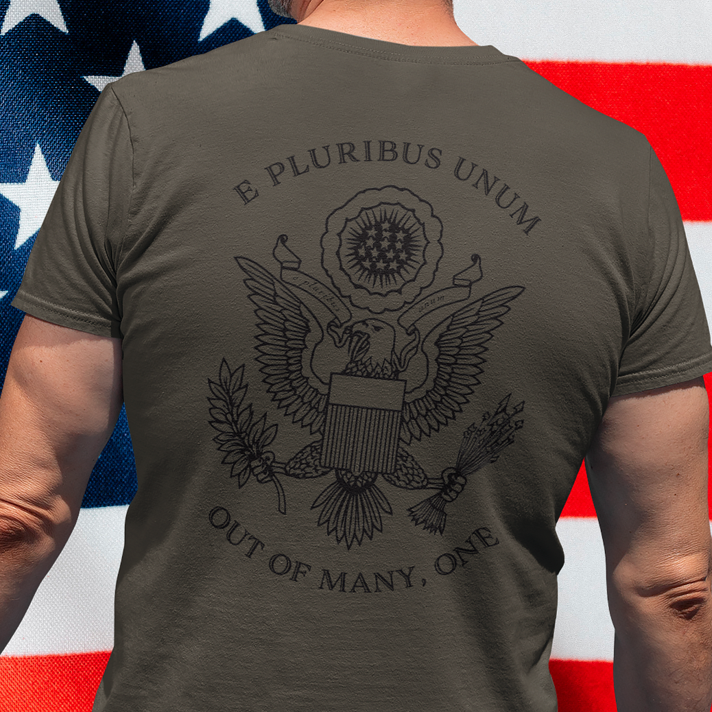 rear view of a man standing in front of an American flag wearing a army green colored fabric patriotic shirt with an americantees.us logo on front and the great seal of the united states on the back 