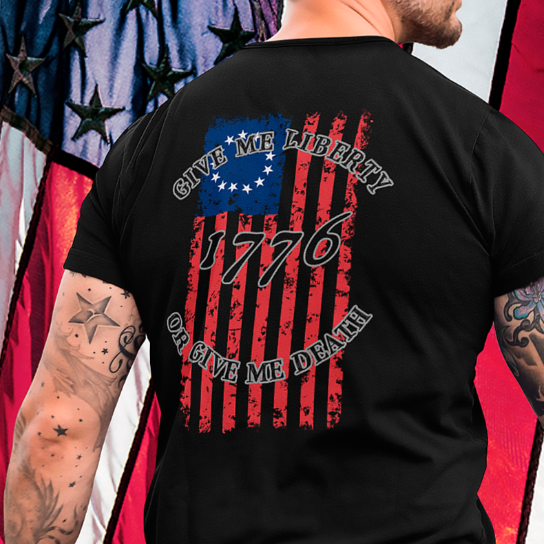 rear view of a man with tattoos and wearing a black colored fabric shirt with americantees.us logo on front and betsy ross flag with the phrase give me liberty or give me death and 1776 on the back