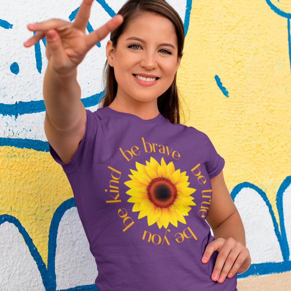 be-true-be-you-be-kind-be-brave-team-purple-t-shirt-sunflower-womens-shirt-mockup-of-a-woman-doing-the-peace-sign-against-a-street-art-wall