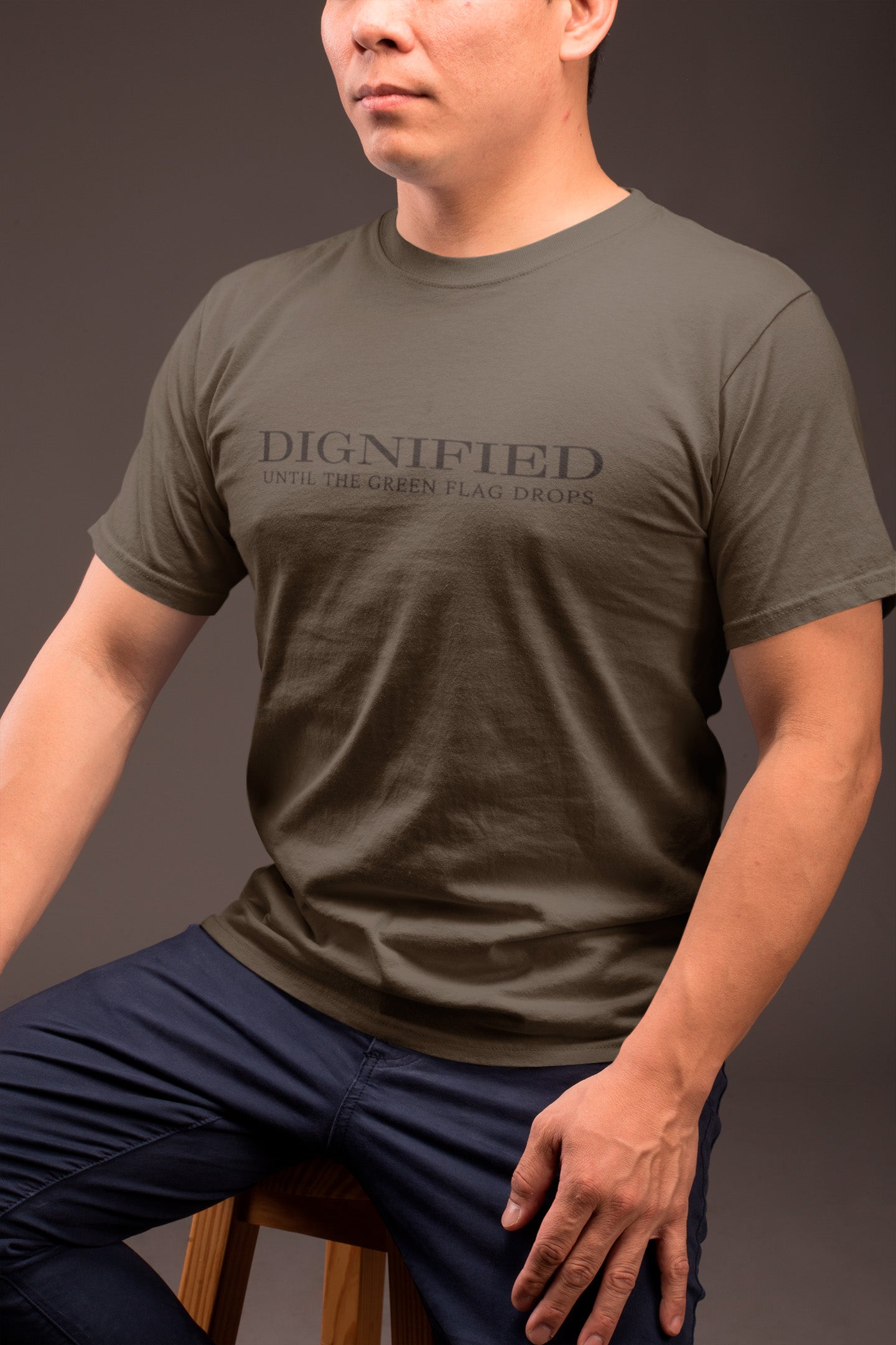 dignified-until-the-green-flag-drops-army-green-mens-t-shirt-crew-neck-tee-mockup-of-a-guy-in-blue-trousers-sitting-on-a-stool-against-a-dark-gray-wall