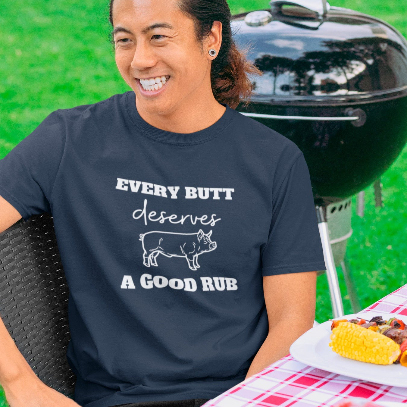 every-butt-deserves-a-good-rub-navy-blue-t-shirt-with-pig-graphic-barbeque-cooking-unisex-crewneck-tee-mockup-featuring-a-smiling-man-by-a-grill-at-a-bbq-party