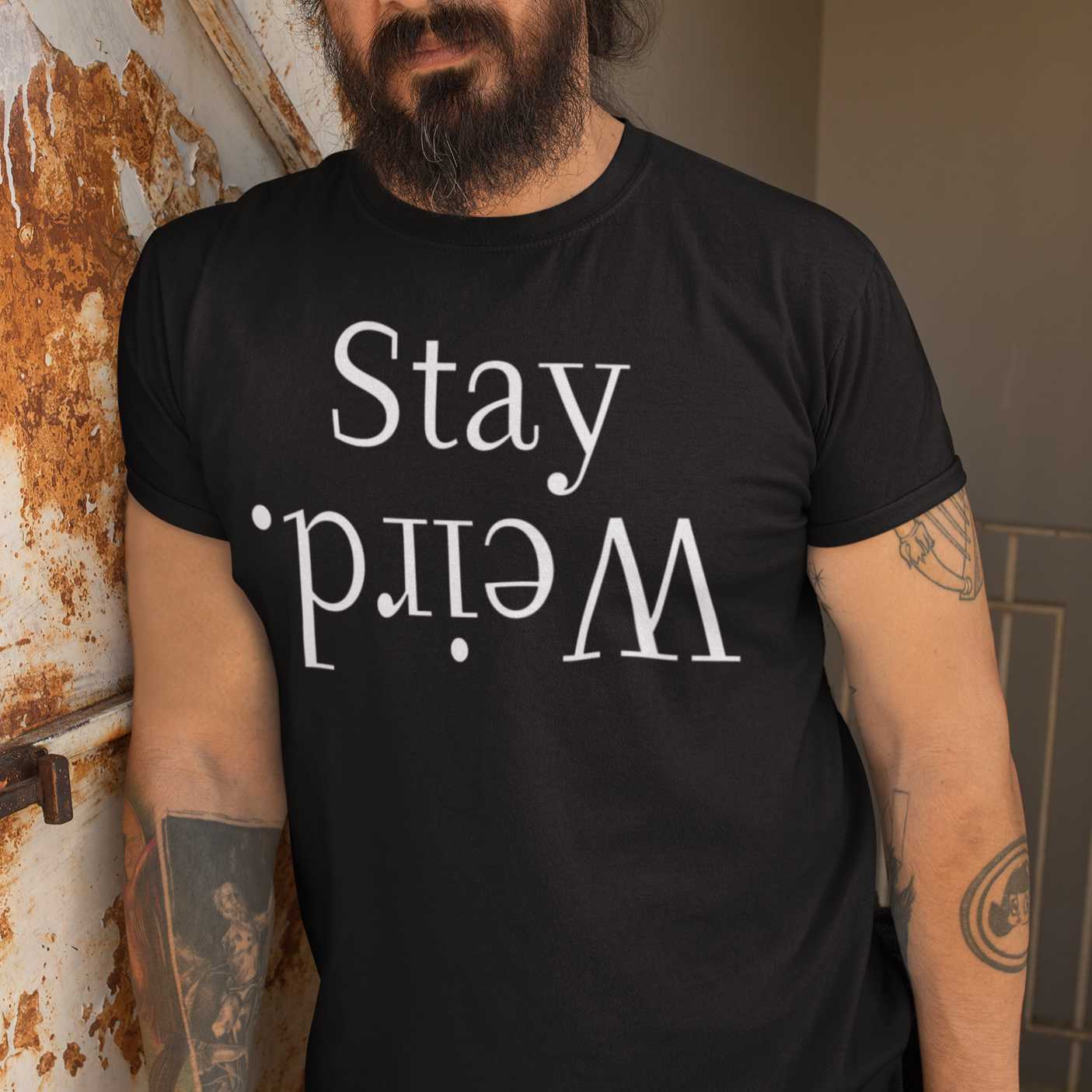 stay-weird-black-t-shirt-unisex-mockup-featuring-a-bearded-man-leaning-against-a-rusty-wall